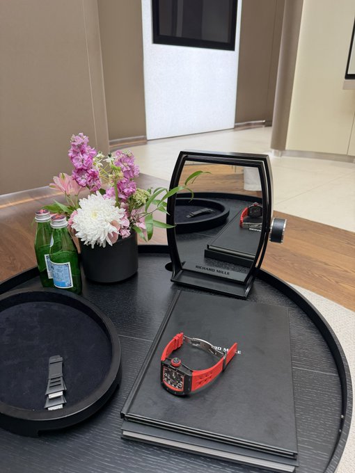 A red Richard Mille RM030 watch is displayed on a black tray alongside a floral arrangement and bottled water. The caption mentions selling this limited edition timepiece for $350,000 to buy more Bitcoin.