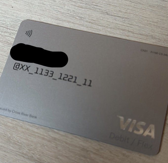 A photograph of a metallic grey Visa Debit Flex card from Cross River Bank lying on a wooden surface, showing the user '@xX_1133_1221_11' with the card number partially redacted in black.