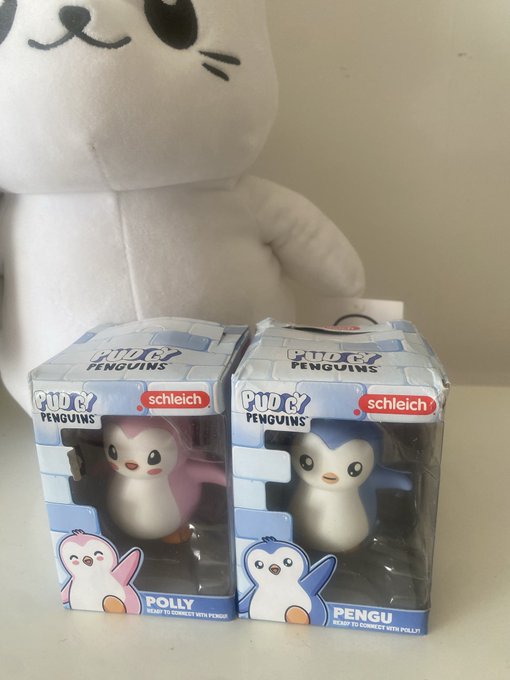 Two boxed Schleich "Pudgy Penguins" figurines, labeled "POLLY" and "PENGU", sit in front of a white plush toy.