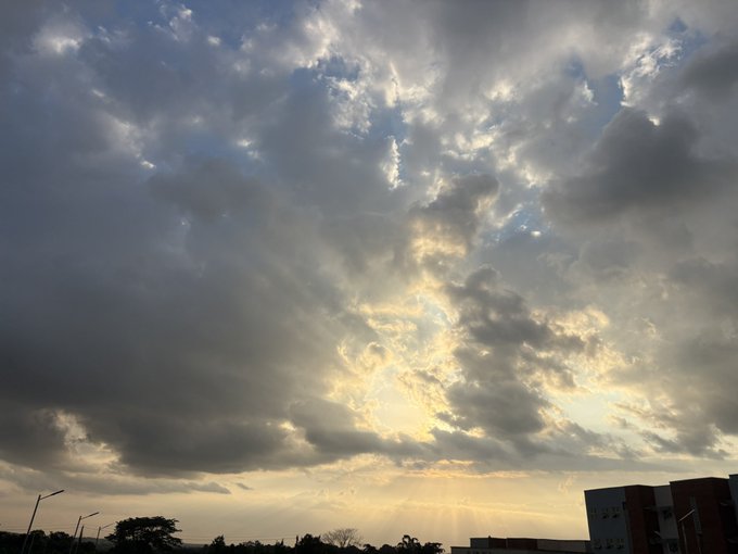 A dramatic sky filled with textured clouds with sun rays breaking through, above a silhouetted landscape with buildings and trees on the horizon.