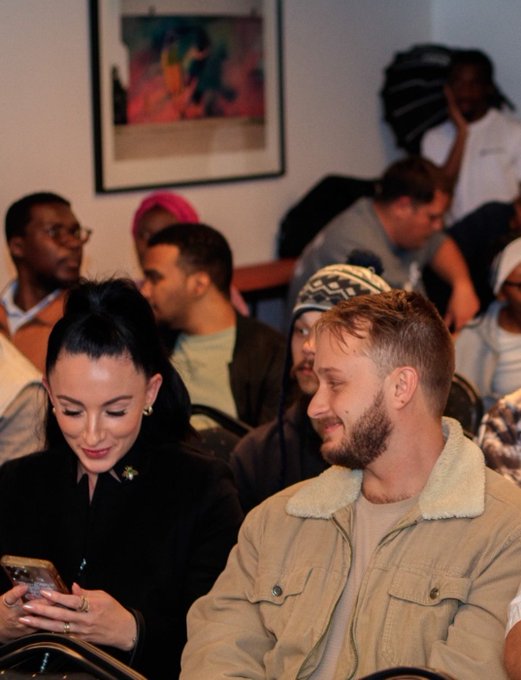 The image shows a group of people sitting down, with one person looking at their phone while another person looks up from their phone. The background includes a painting hanging on the wall behind them. There are several other people present as well, some of whom appear to be engaged in conversation or focused on something else. A caption that reads "I can’t take most web3 events seriously" is also present in the image.