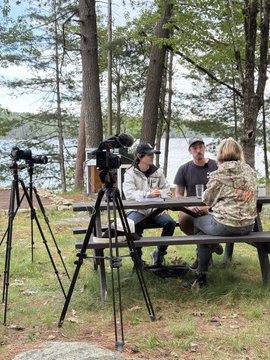 The image shows a group of people sitting around a picnic table, enjoying each other's company outdoors near a body of water. There are two individuals seated at the table with one person standing nearby. The scene appears to be set up for an outdoor event or gathering, possibly related to Bitcoin or cryptocurrency. A camera is positioned on the ground next to the group, suggesting that they might have been recording the event or capturing memories from their time together.