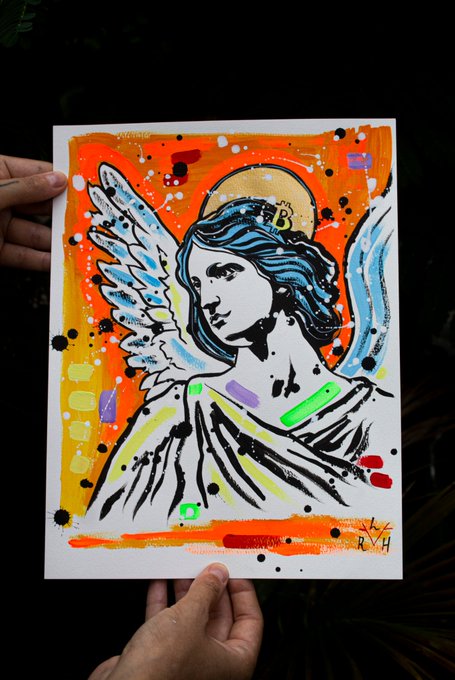 Hands hold a mixed-media painting of a blue-haired angel with a Bitcoin halo against an orange background.