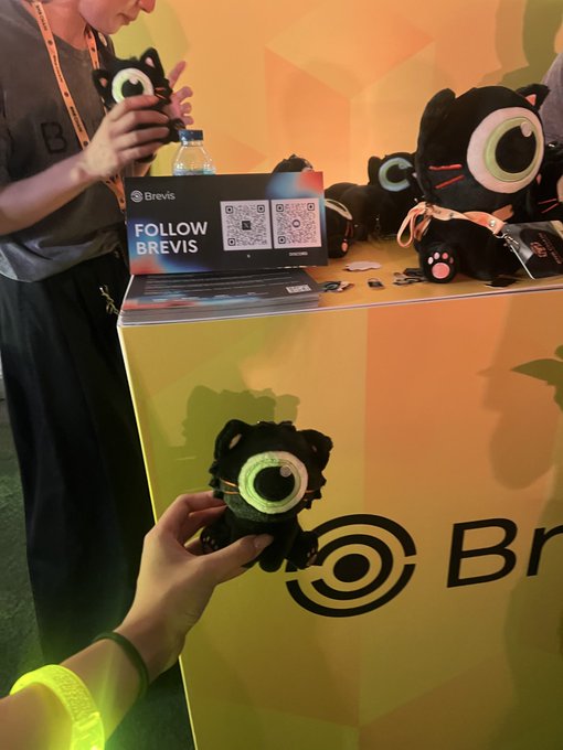 A person holds a black plush toy with a large green and pink eye at a 'Brevis' booth, with more plush toys and a laptop displaying a QR code and 'FOLLOW BREVIS' visible on the orange and yellow table.