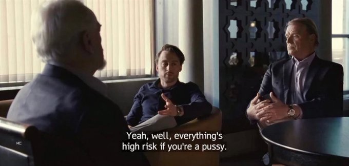 A scene from a film with three men sitting in a room, with subtitles reading 'Yeah, well, everything's high risk if you're a pussy.'