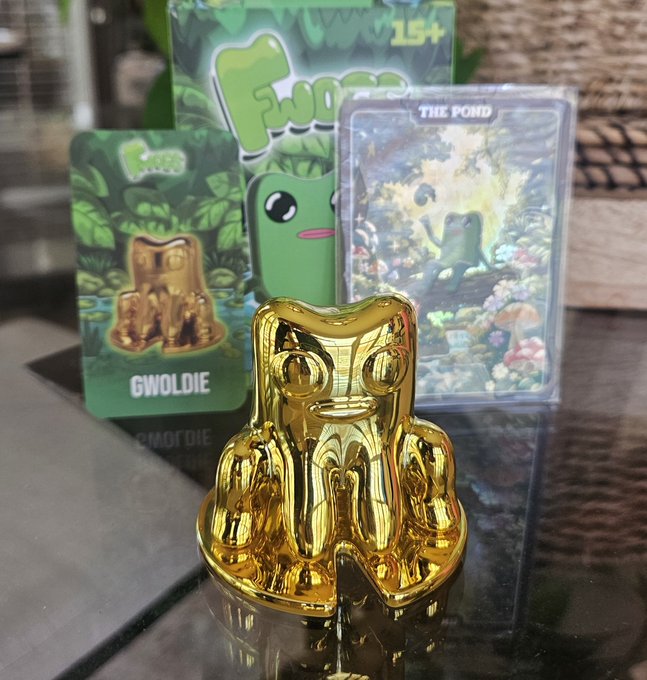 A shiny gold "Gwoldie" figurine sits on a reflective surface in front of its green jungle-themed Fwog blind box packaging (marked 15+) and an illustrated trading card labeled "THE POND," matching the poster's excitement about pulling the rare gold variant from a single blind box.