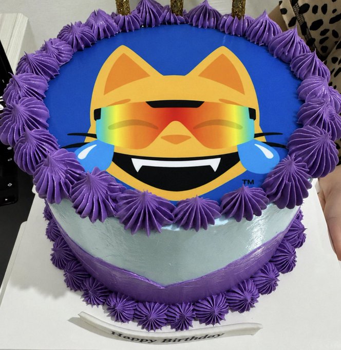 A birthday cake decorated with purple frosting rosettes and a laughing cat emoji wearing rainbow sunglasses on top, matching the celebratory "Happy birthday to me" post with a "Happy Birthday" banner at the base.