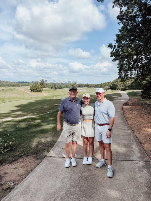 Three people posing together on a paved path at a golf course with a green fairway and city skyline visible in the distance.