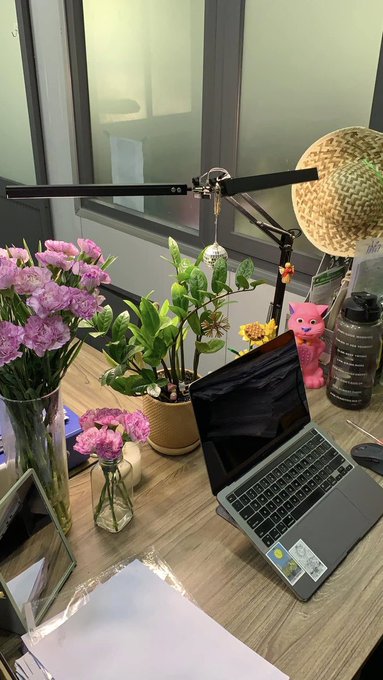 A desk setup featuring a laptop, a black monitor lamp, pink and yellow flowers in vases, a small potted plant, a pink figurine, and a straw hat.