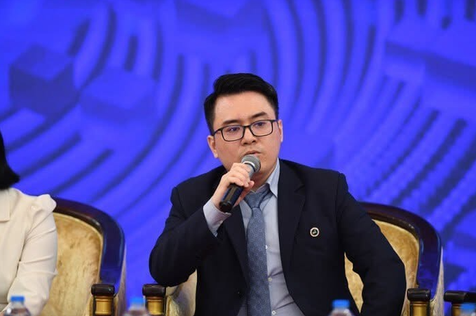 A man wearing a dark jacket, glasses, and tie speaking into a microphone while seated on a stage with a blue patterned backdrop during what appears to be a panel discussion.
