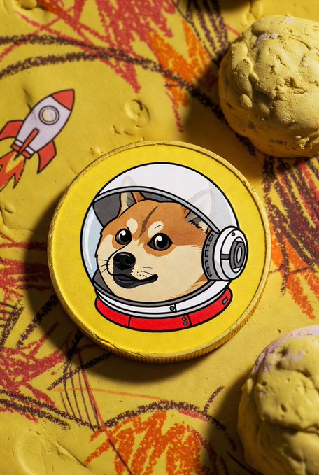 A round yellow enamel pin depicting a Shiba Inu dog wearing a space helmet, lying on a yellow textured surface with a rocket ship drawing and two cookies nearby.