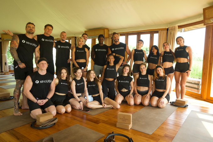 A group of people wearing black "strcUSX" branded clothing pose together on yoga mats in a wooden-floored studio.