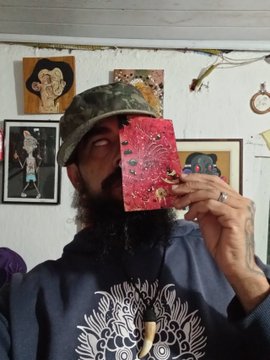 A bearded man wearing a camouflage cap and a dark hoodie with a white graphic, holding a red ornate mask over his face, with framed artwork visible on the wall behind him.