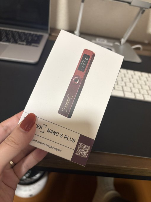 A hand holds a white box for a 'Ledger Nano S Plus' crypto signer, with a laptop visible in the background.