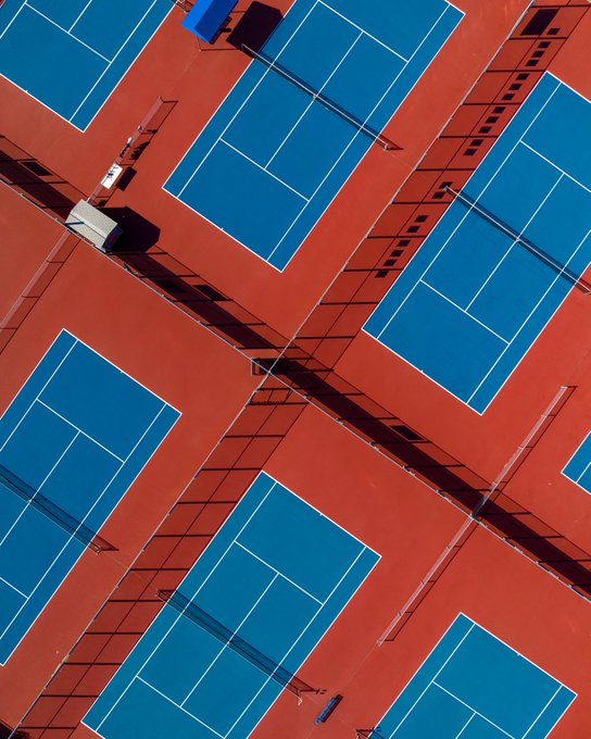 An aerial view of multiple blue tennis courts surrounded by reddish-brown pathways with shadows cast by fences.