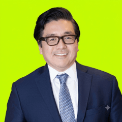 A professional headshot shows a smiling man with dark hair, wearing black-framed glasses, a navy suit, white shirt, and patterned blue tie, set against a bright green background. The image accompanies a post about Tom Lee's Bitmine (BMNR) significantly expanding its Ethereum holdings to 6.04 million ETH, with the pictured individual likely being Tom Lee, the founder associated with the company's crypto treasury strategy.