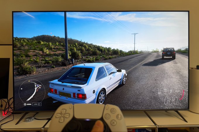 A person holds a white game controller while playing a racing video game on a large television, perfectly illustrating the idea of "How to Kick Off the Weekend" with some gaming. The screen displays a white car driving on a road with a "Fortress" speedometer visible in the corner.