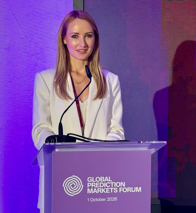 A blonde woman in a white blazer speaks at a podium for the 'Global Prediction Markets Forum' dated '1 October 2026' against a purple backdrop.