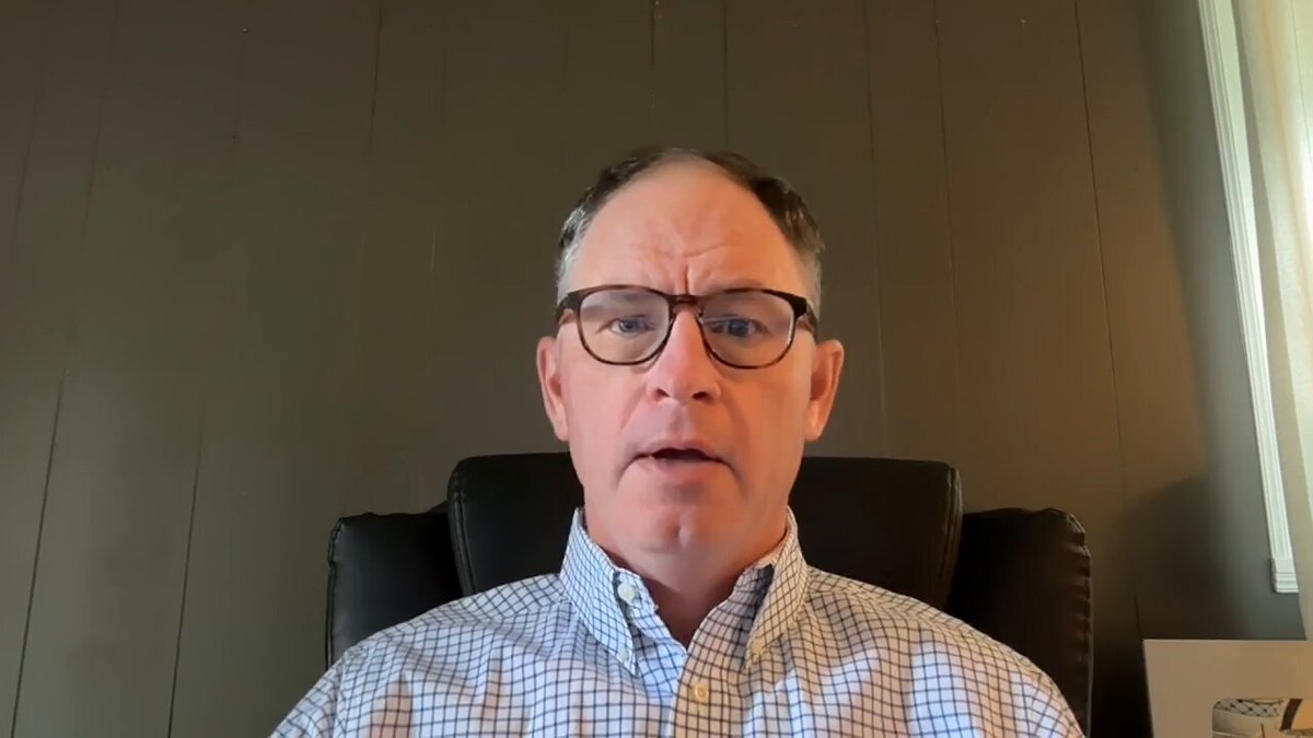 The image shows a man wearing glasses sitting in an office chair, with his face turned towards the camera. He appears to be giving a presentation or speaking about something important. The background of the image features a gray wall, which complements the man's attire. There is also a book visible on the right side of the image.