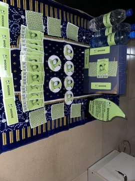 A blue table covered with a patterned cloth displays various green and white Baboon brand product packages, boxes, and pouches, likely soaps and teas, with a water bottle at the top.