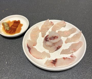 A white plate of raw fish sashimi, with slices arranged in a flower pattern around a center piece, accompanied by a small dish of soy sauce, wasabi, and pickled ginger.