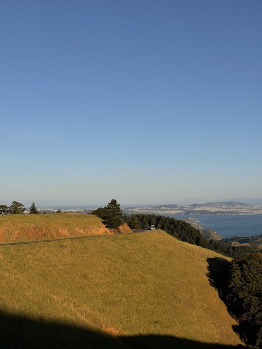 A scenic view from a grassy hillside overlooks a coastal city and bay under a clear blue sky, with a paved road winding through the rolling green terrain.