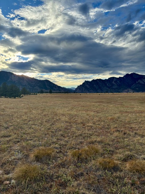 A scenic landscape featuring a vast grassy plain with rocky mountains in the background under a dramatic cloudy sky at sunset.