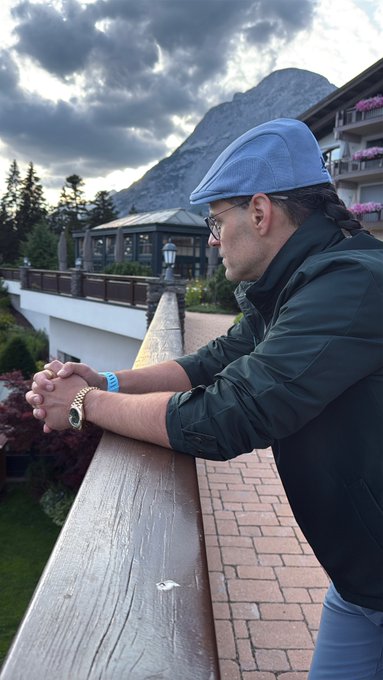 A man wearing a flat cap, glasses, and a dark jacket stands at a wooden balcony railing with hands clasped, overlooking a mountain landscape with dramatic clouds and a resort-style building.