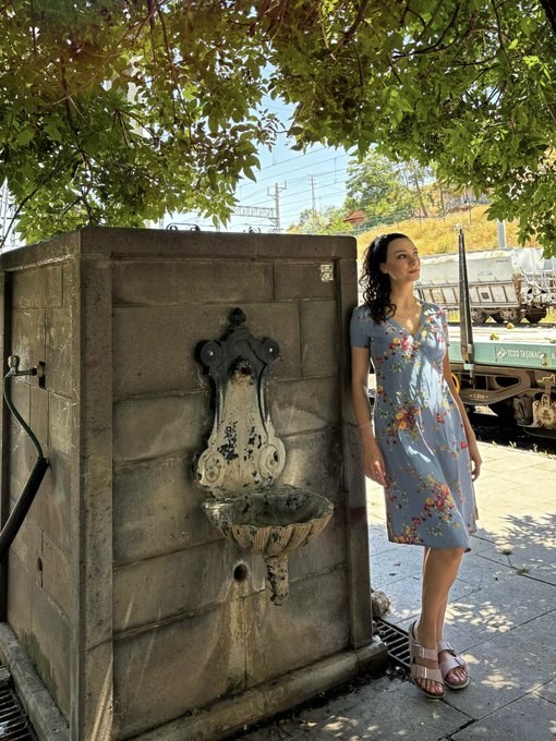 A woman in a floral dress leans against an ornate stone fountain under a leafy canopy, embodying a serene "Good Morning" moment. The visible text "SOTO SHIPPING" on a truck in the background adds an urban touch to the peaceful Thursday vibe.