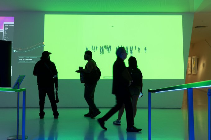 Silhouetted visitors walking past a large green-lit digital display in an art gallery or museum, with a smaller screen showing information on the left.