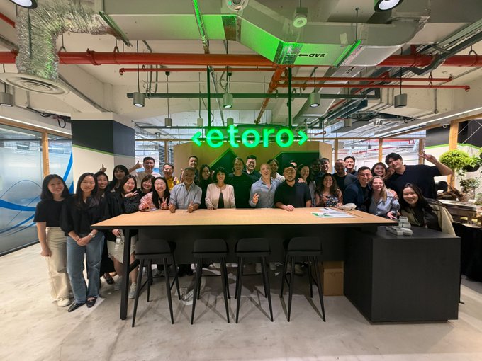 A group of around 20 people pose for a team photo inside the eToro office in Singapore, gathered around a large wooden table beneath the company's logo displayed on the wall. The image captures the welcoming team environment that aligns with the post celebrating a visit to eToro's Singapore office.
