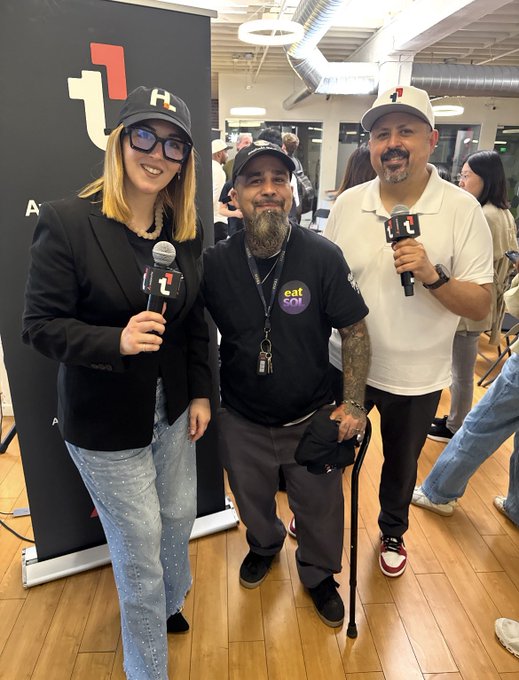 Three people smiling for a photo at an event, with two holding microphones bearing a '+1' logo; one wears a black shirt that reads 'eat soi'.
