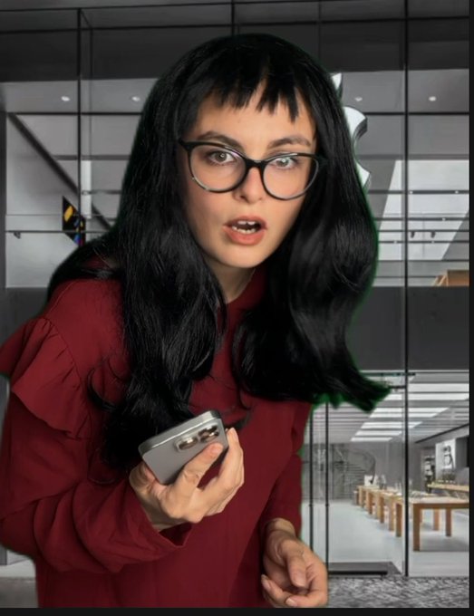 A woman with black hair, glasses, and a maroon top looks surprised while holding a smartphone in what appears to be an Apple Store.
