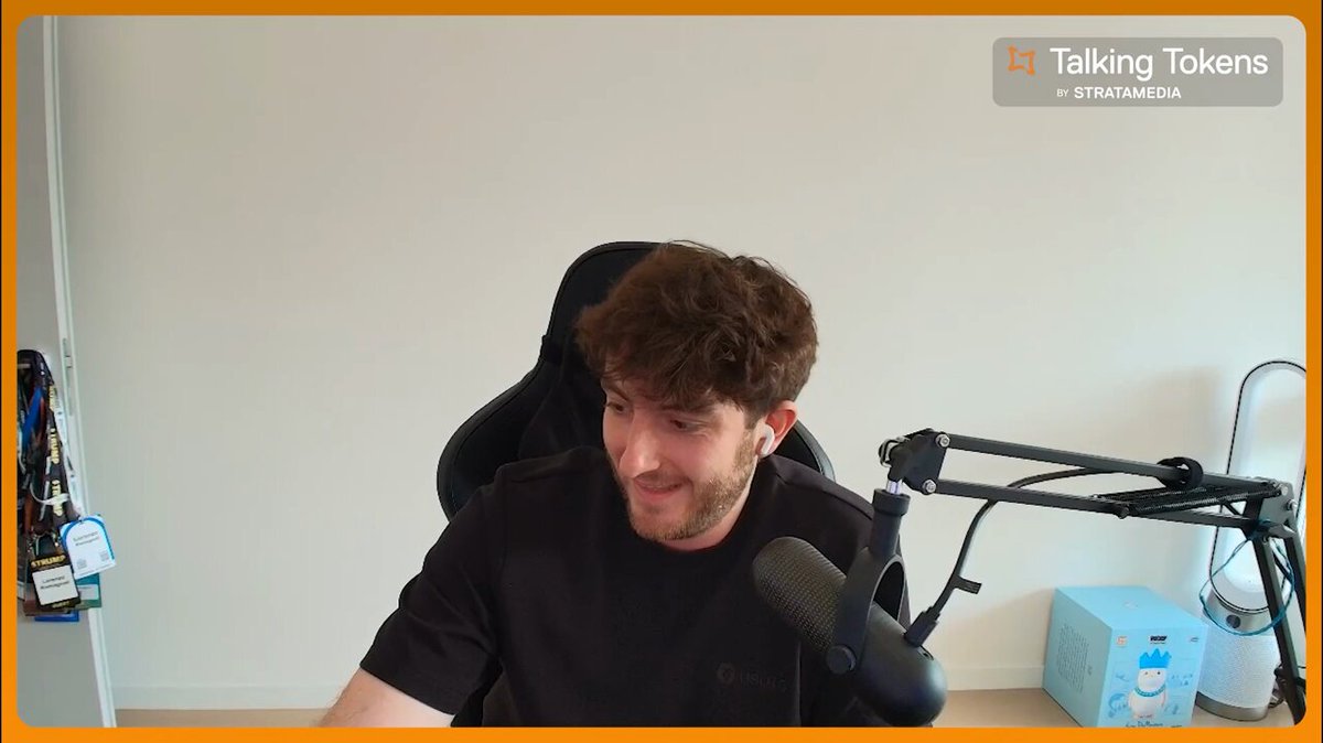 The image shows a man sitting at a desk using a microphone, likely for a podcast or live stream. The background features a white wall and an orange frame with text that reads "Stablecoins are freedom in the form of code." This suggests that the man might be discussing the concept of stablecoins as a means to protect one's assets from inflation or currency fluctuations.