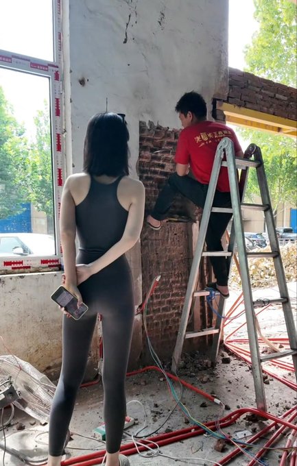 A woman in a black outfit stands holding a phone while watching a man in a red shirt on a ladder working on an exposed brick wall at a construction site.