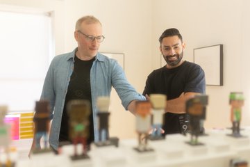 Two men look down at a table covered with small colorful product displays or prototypes in a bright indoor workspace.