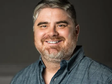 A professional headshot of a smiling man with short graying hair and a beard, wearing a blue button-down shirt against a gray background.