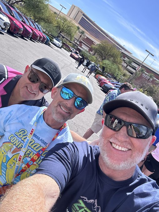 A selfie shows three men wearing sunglasses, baseball caps, and colorful t-shirts smiling together in an outdoor parking lot setting.