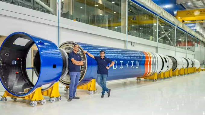 Two Skyroot Aerospace engineers pose beside a large blue rocket stage in a manufacturing facility, with Japanese text visible on the rocket's fuselage, illustrating the company's hardware capabilities as it reportedly nears raising up to $300 million from Fidelity, General Catalyst, and Benchmark.
