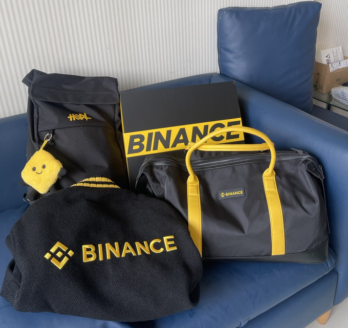 A collection of black and yellow Binance-branded merchandise, including a backpack, a soft hat, a box, and a duffel bag, sits on a blue couch.
