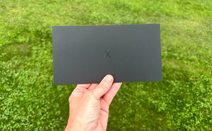 A hand holds a minimalist dark gray envelope featuring a subtle 'X' logo, photographed against a grass background.