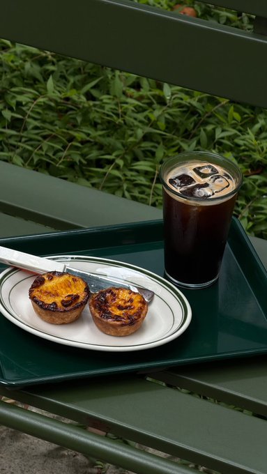 A green tray on a bench holding two Portuguese egg tarts (pastéis de nata) and a glass of iced coffee, with green plants in the background.