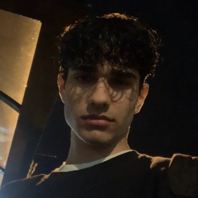 A young man with dark curly hair takes a moody nighttime selfie, casting an intense, contemplative gaze toward the camera. The dimly lit setting and serious expression align with the post's confident, self-assured tone of a 22-year-old professional trader seeking connections.