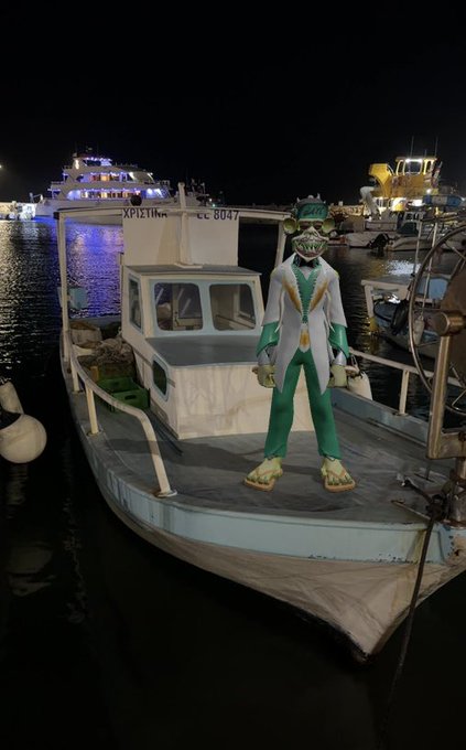 A green lizard/iguana character wearing a white suit with gold trim standing on the deck of a fishing boat at a harbor at night, with other boats visible in the background.