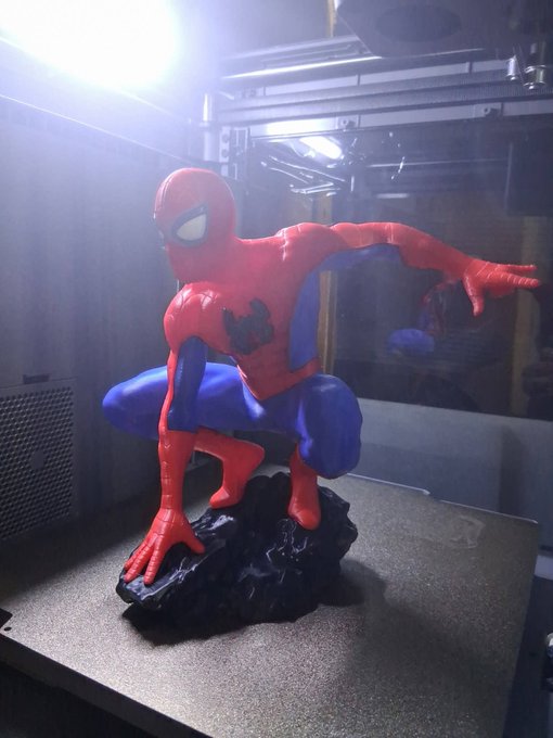 3D-printed Spider-Man figurine in a crouching pose on a black rock base, displayed inside what appears to be a 3D printer enclosure.