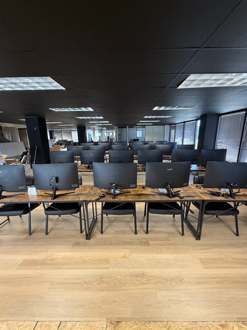 A modern computer classroom with rows of black monitors and chairs arranged on wooden desks, featuring a black ceiling with fluorescent lighting and large windows along the back wall.