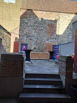 An outdoor ICP meetup venue in Ghent, featuring stone and brick walls with two blue "Internet Computer" branded banners displayed near a small stage area, where the poster gathered with community members including @ZeroFuxBJ.
