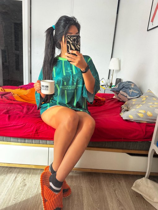 A young woman with pigtails sitting on a bed with red sheets, wearing an oversized teal and green striped jersey, holding a mug, with her phone obscuring her face as she takes a mirror selfie.