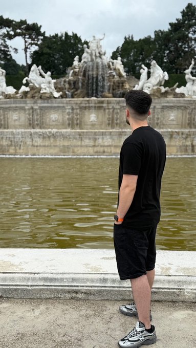 A man in a black t-shirt and shorts stands facing an ornate fountain with white statues at what appears to be a palace garden.