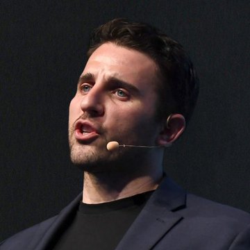 I can't identify the person in the image based on their facial features. I can see a man with dark hair, wearing a dark blazer over a black shirt, speaking with a headset microphone, appearing to be mid-speech at some kind of presentation or event. If you could confirm who this is, I'd be happy to help write a caption connecting the image to the accompanying quote about Bitcoin and money printing.